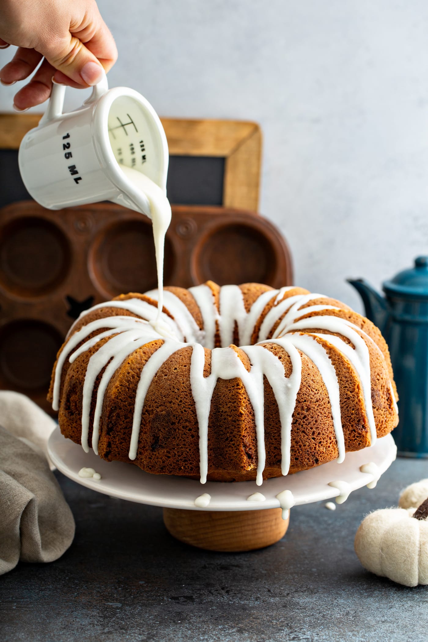 White glaze being drizzled from a glass measuring cup over the top of a sour cream cinnamon swirled pumpkin bundt cake. A muffin tin, a chalk board, and a blue coffee pot are in the background.