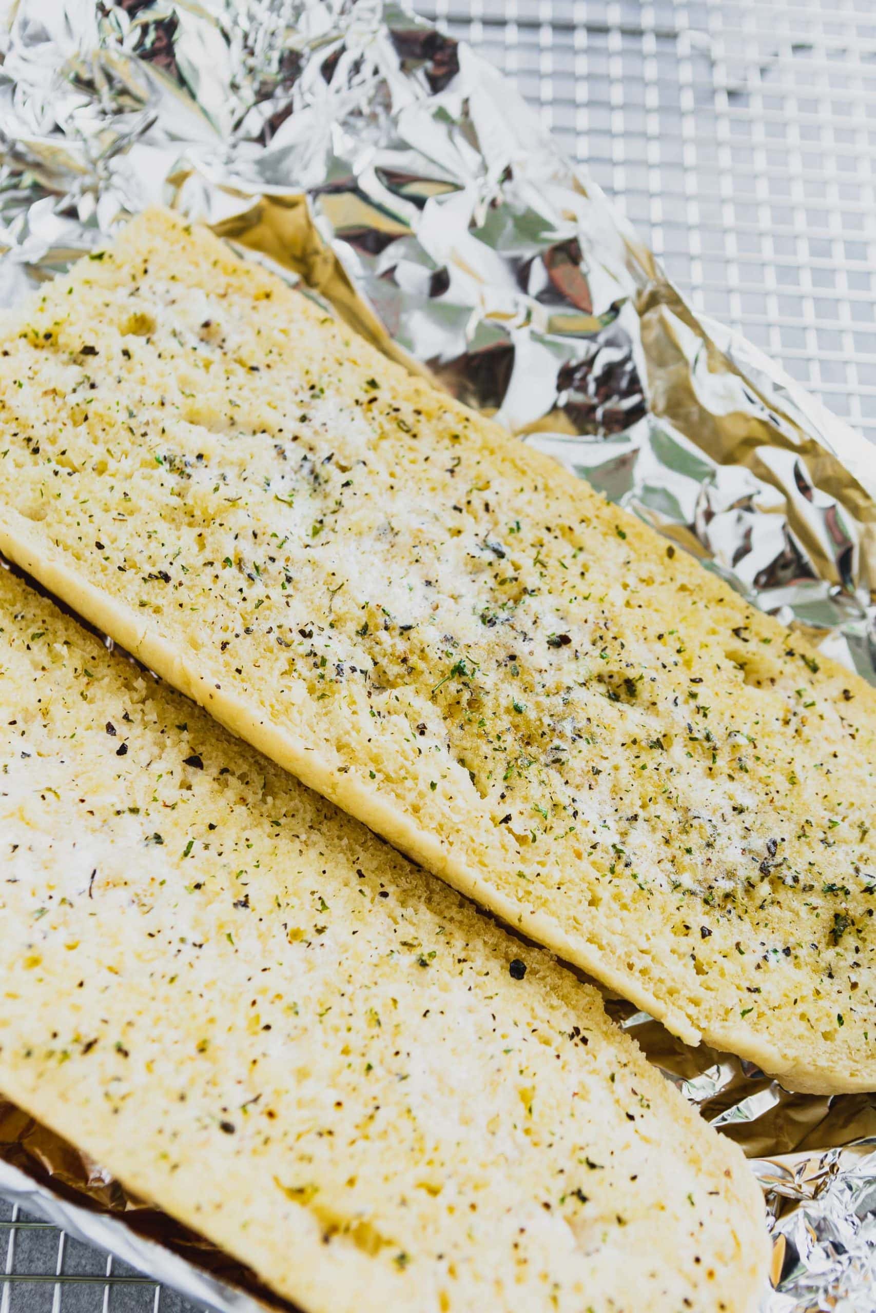 a photo of a loaf of french bread that has been cut in half with both halves facing up that have been covered in butter, garlic and chopped parsley. the loaves are sitting on a large sheet of aluminum foil.