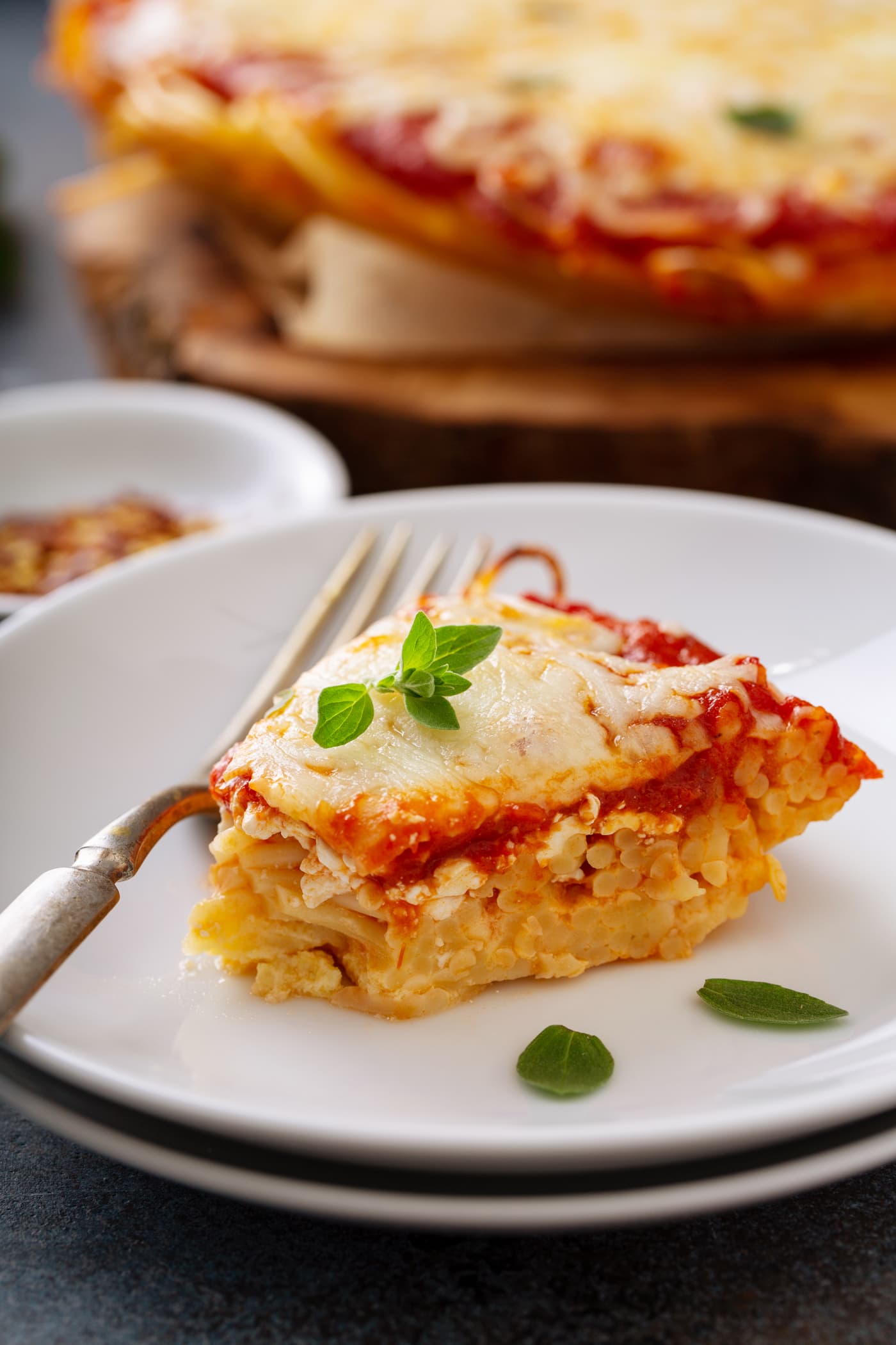 A slice of baked spaghetti pie on a white dinner plate. A fork is next to the spaghetti pie and a few basil leaves are scattered on the pie and the plate. A small container of red pepper flakes and the rest of the spaghetti pie are in the background.