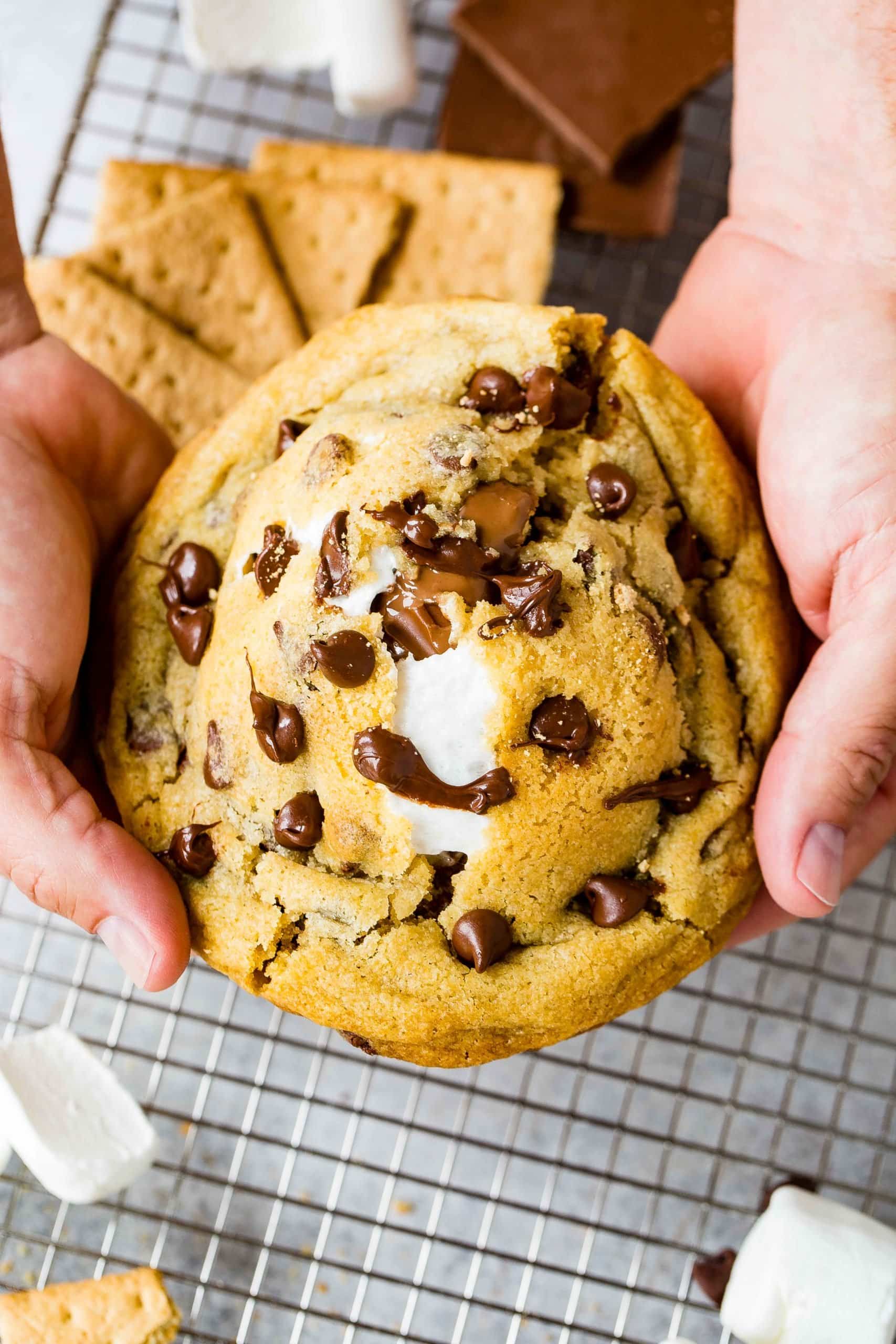 A s'mores stuffed chocolate chip cookie being broken in half exposing melted marshmallow inside. The large cookie is packed with chocolate chips and marshmallow and graham crackers. Graham crackers are on the cooling rack below the cookie.