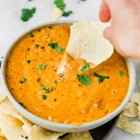 A bowl of chilis queso dip. It is cheesy and thick with a few parsley flakes scattered on top. A tortilla chip is being dipped into the cheese dip.