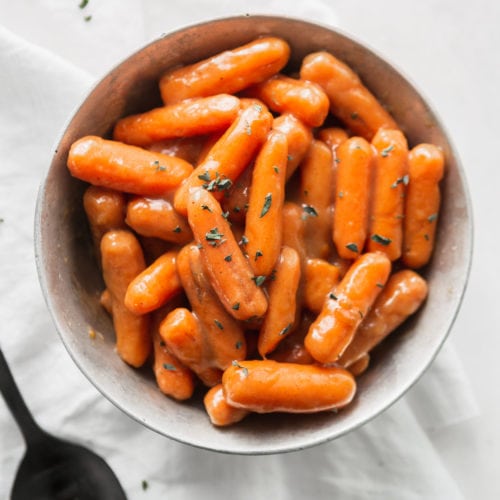 A bowl of orange glazed baby carrots. A serving spoon is next to the bowl.