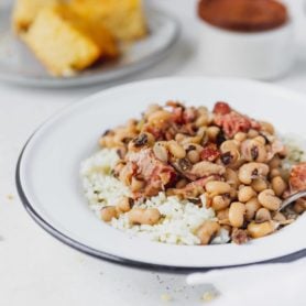 A plate of southern instant pot black eyed peas over rice. There are chunks of ham in the peas and a plate of cornbread in the background.