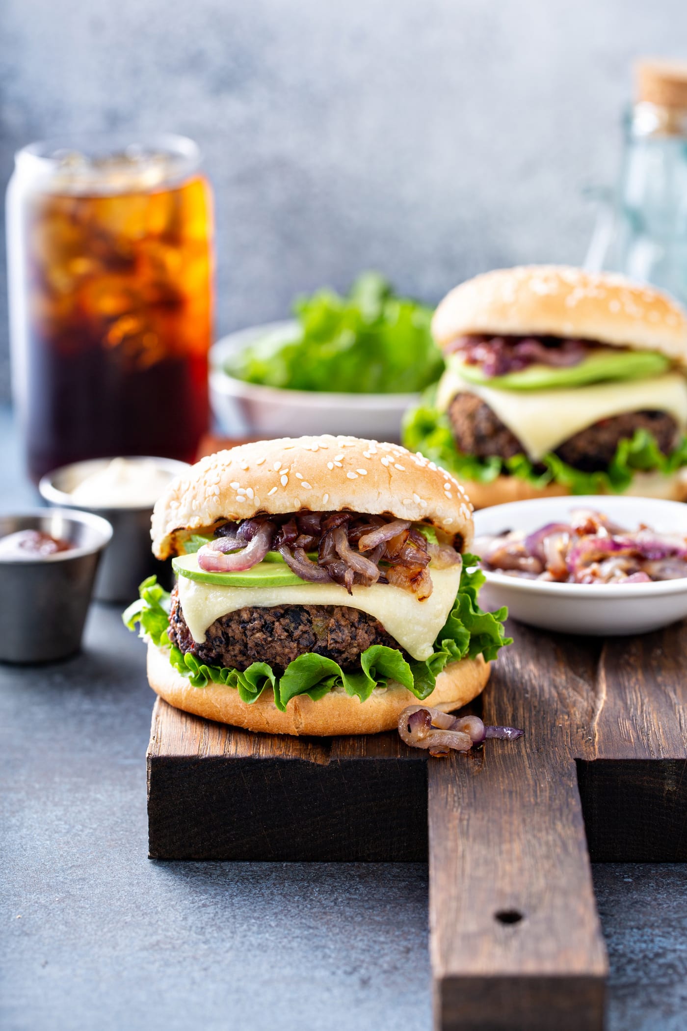A black bean burger on a sesame seed bun topped with onions, avocado, cheese and lettuce. There is another burger, a bowl of lettuce, a container of onions, a container of mayo and a glass of iced soda in the background.