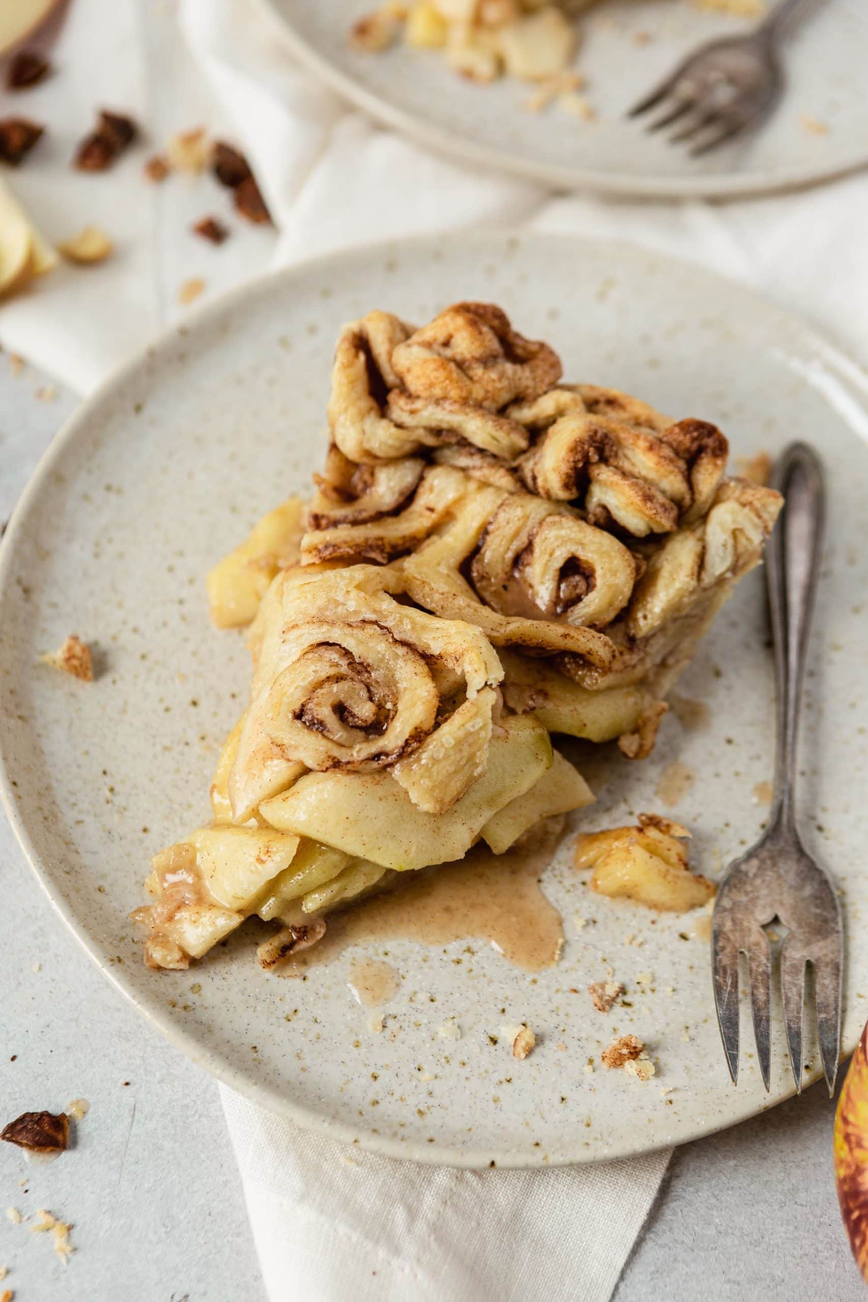 A slice of cinnamon roll apple pie. The Apple are juicy and the cinnamon roll top crust is flaky and beautiful. A fork is resting on the plate next to the pie.