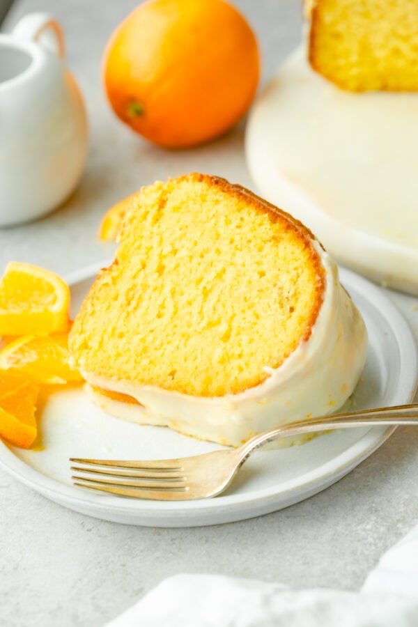 a photo of a slice of bright orange bundt cake topped with a white glaze sitting on a white plate with a fork in front of if and small orange slices scattered around it.
