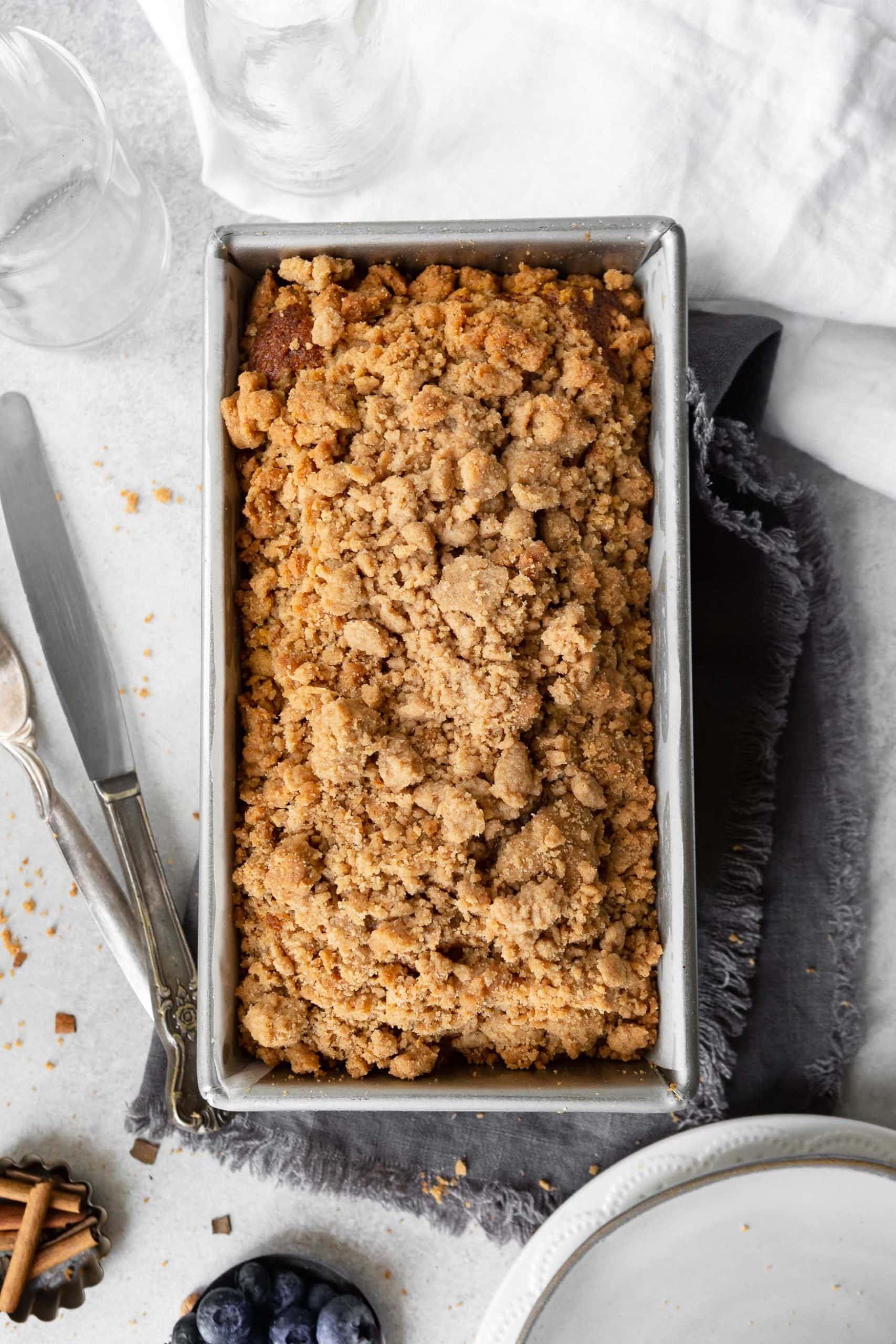 a photo of a loaf of pumpkin bread topped with a cinnamon crumb topping in a metal loaf pan.