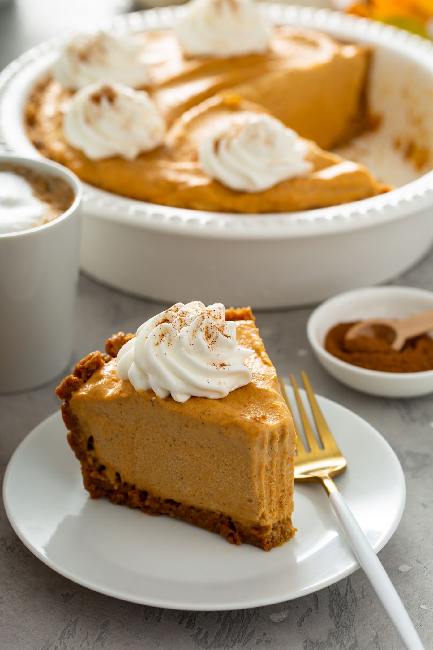 A slice of pumpkin ice cream pie on a dessert plate with a fork beside it. There is a dollop of whipped cream sprinkled with cinnamon on top of the slice of pie and a bite has been taken out of the pie. A bowl of cinnamon with a wooden spoon, a white mug of hot cocoa topped with whipped cream and a white pie dish with pumpkin ice cream pie is in the background.