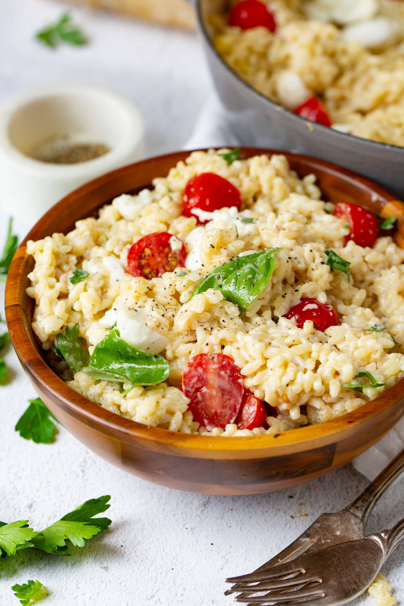 A serving bowl of caprese orzo salad. The salad contains tomatoes, mozzarella chuncks and fresh basil. A few basil leaves, tomatoes and a small container of oil are sitting by the salad.