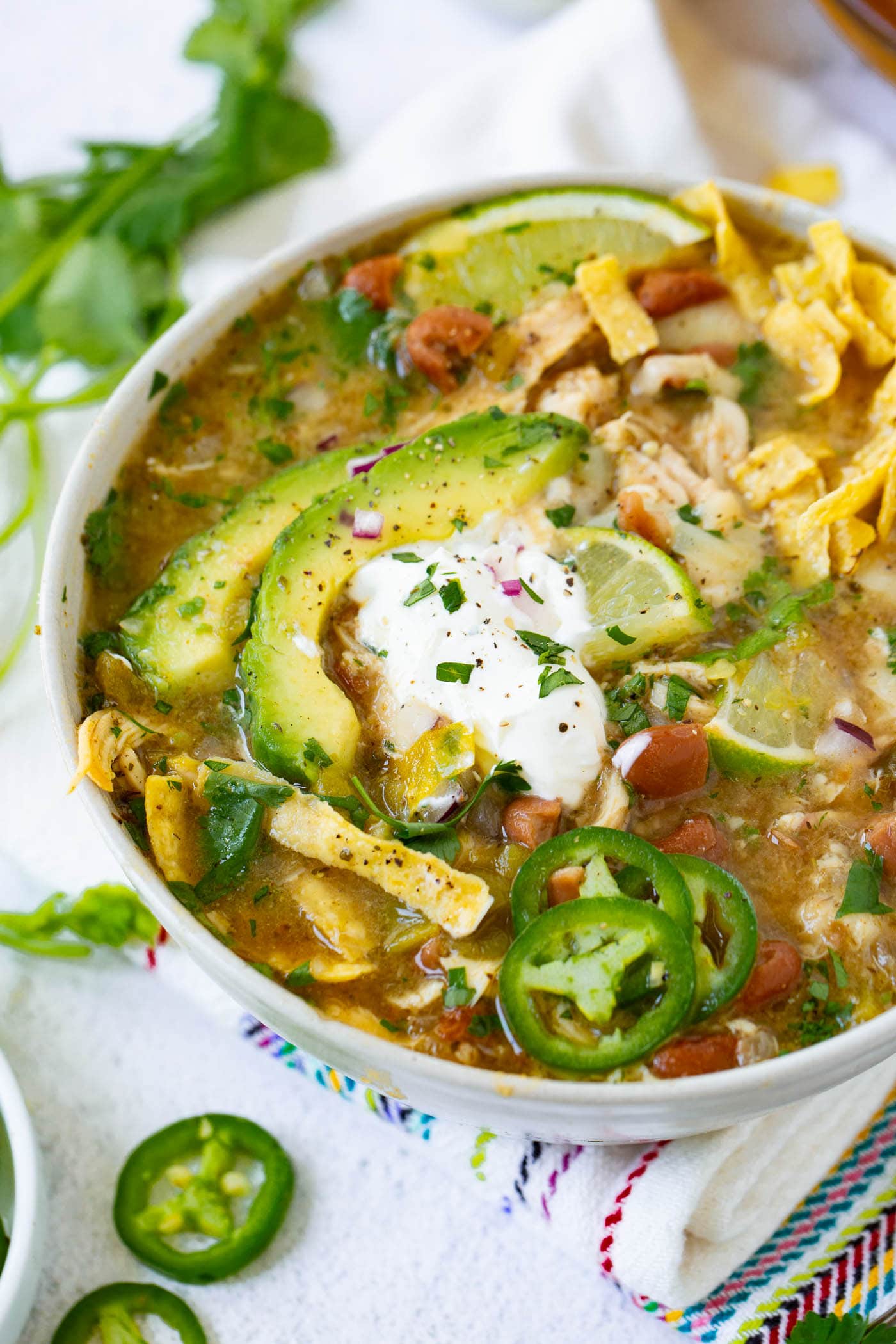 a photo of a bowl full of white chicken chili topped with a dollop of sour cream, avocado slices, tortilla chip strips and slices of fresh jalapeno.