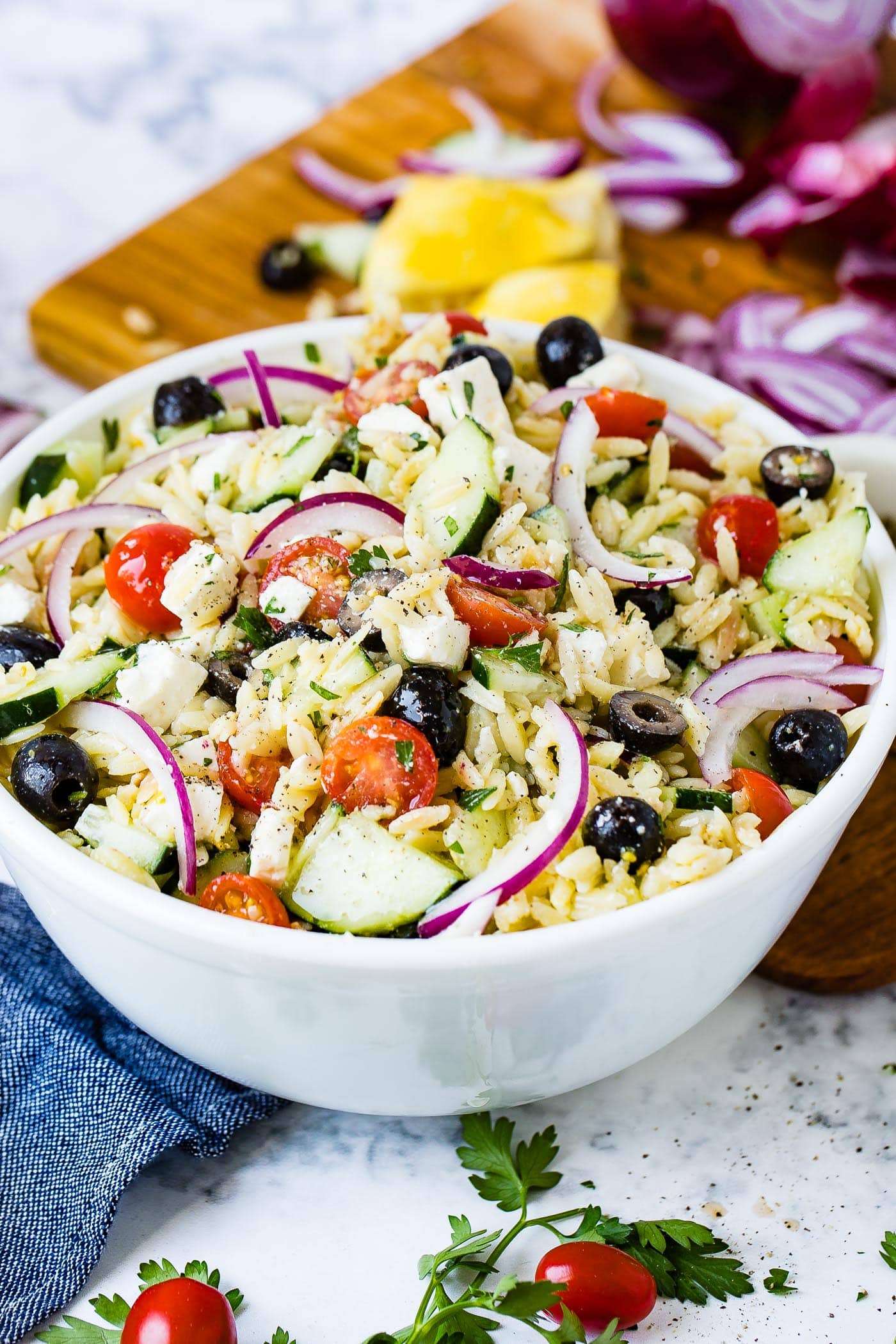 A white bowl full of Greek orzo pasta salad with black olives, cucumbers, cherry tomatoes, slices of red onions and feta cheese.