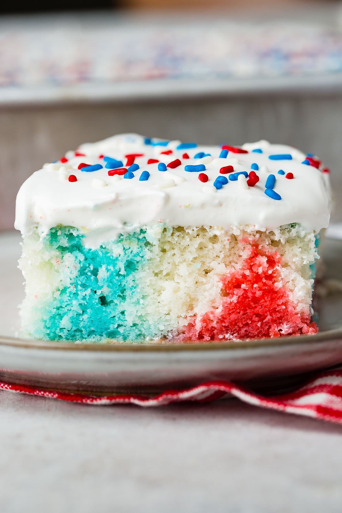 A piece of red, white and blue poke cake on a dessert plate. The cake is frosted with white frosting and sprinkled with red, white and blue sprinkles.