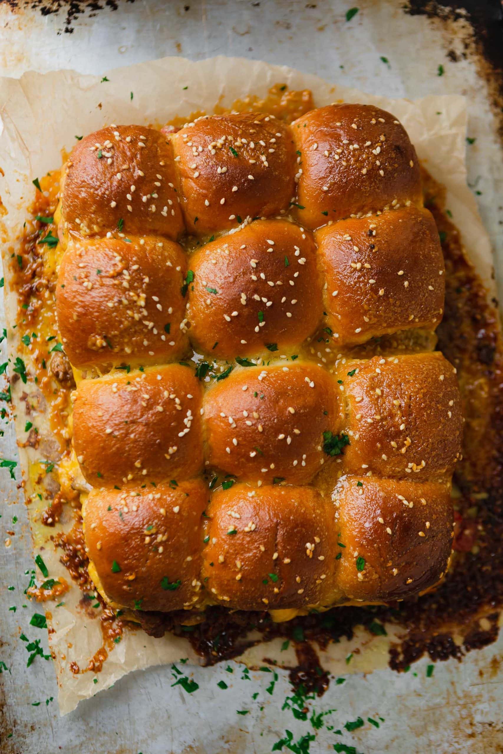 A panful of cheeseburger sliders. There is melted cheese oozing from the rolls that are sprinkled with sesame seeds.
