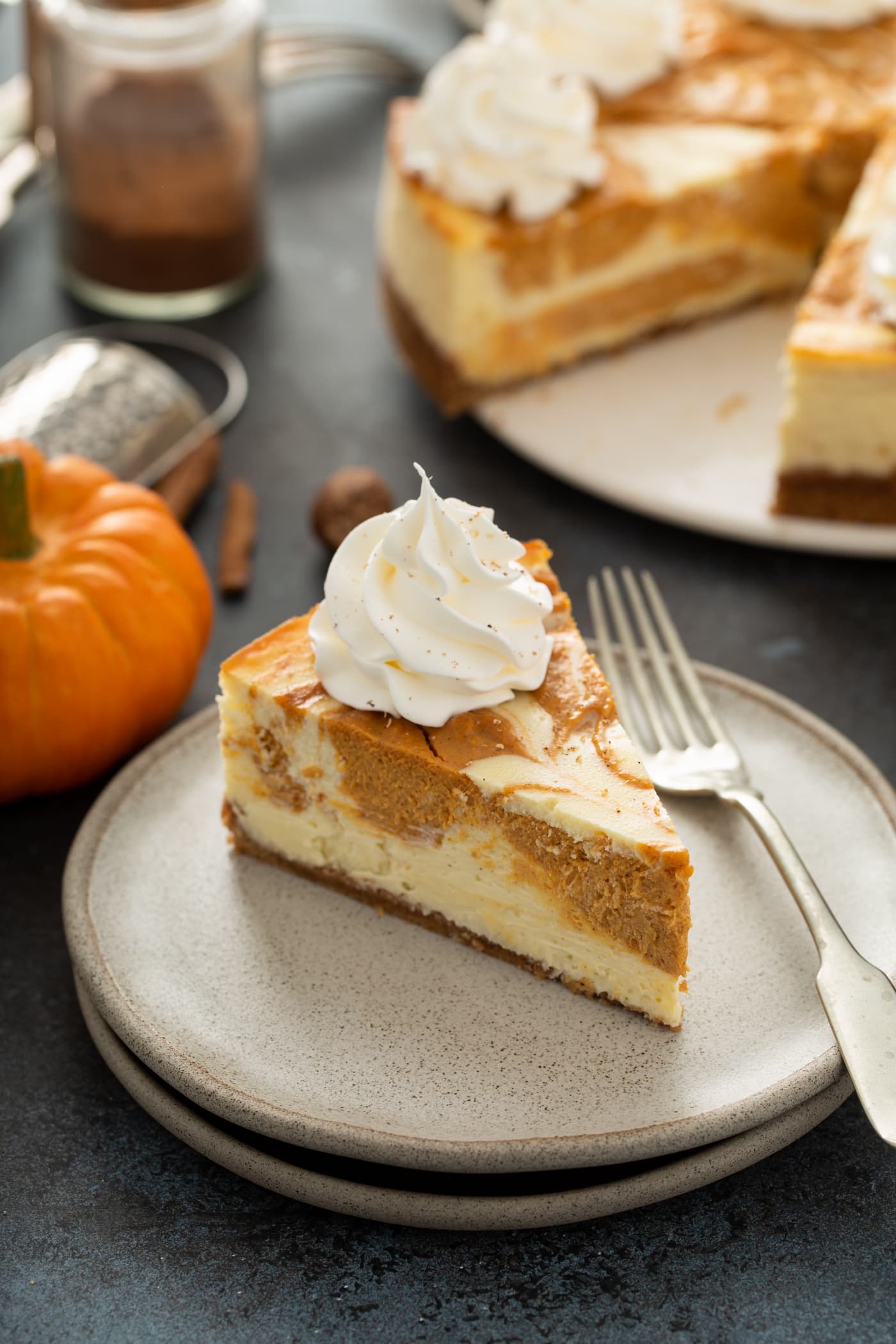 A slice of swirled pumpkin cheesecake on a dessert plate with a fork. The rest of the cheesecake, and a small pumpkin are in the background.