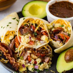 A plate with two apple chicken sausage burritos. The burrito has sausage, eggs, cheese. There is a bowl of salsa and an avocado that is cut in half next to the burritos.