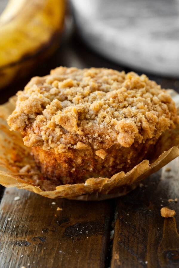 a photo of a banana muffin with crumb topping sitting in a muffin liner that has been removed slightly from the sides of the muffin.