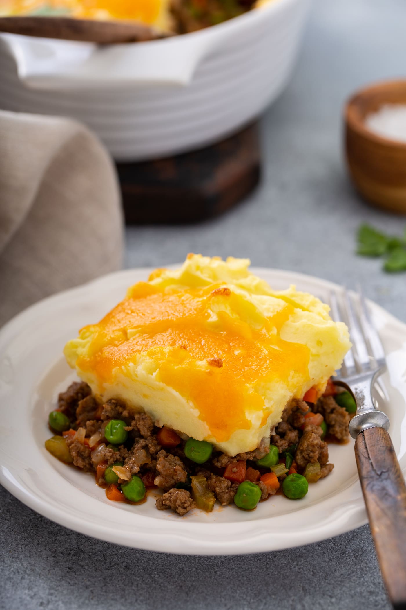A serving of shepherd's pie on a white plate with a fork beside it. There is a napkin and a casserole dish in the background.