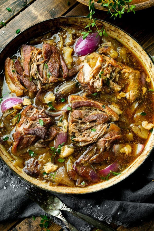 a photo of chunks of braised pork butt in a large dutch oven sitting in a rich sauce full of apple slices, onion slices and fresh herbs.