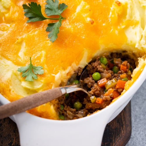 A casserole dish containing shepherd's pie with one spoonful removed and the serving spoon resting in the dish. You can see hamburger, carrots, and peas in the pie and it is topped with mashed potatoes, and melted cheddar cheese. There is a few parsley leaves on top.