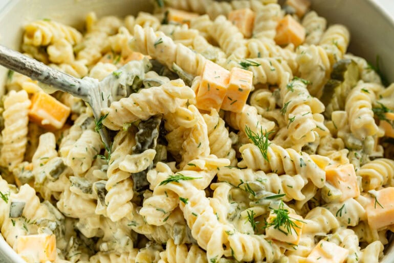 A close up of dill pickle pasta salad with pasta in a creamy sauce, pickles, cheddar cheese, and garnished with dill.