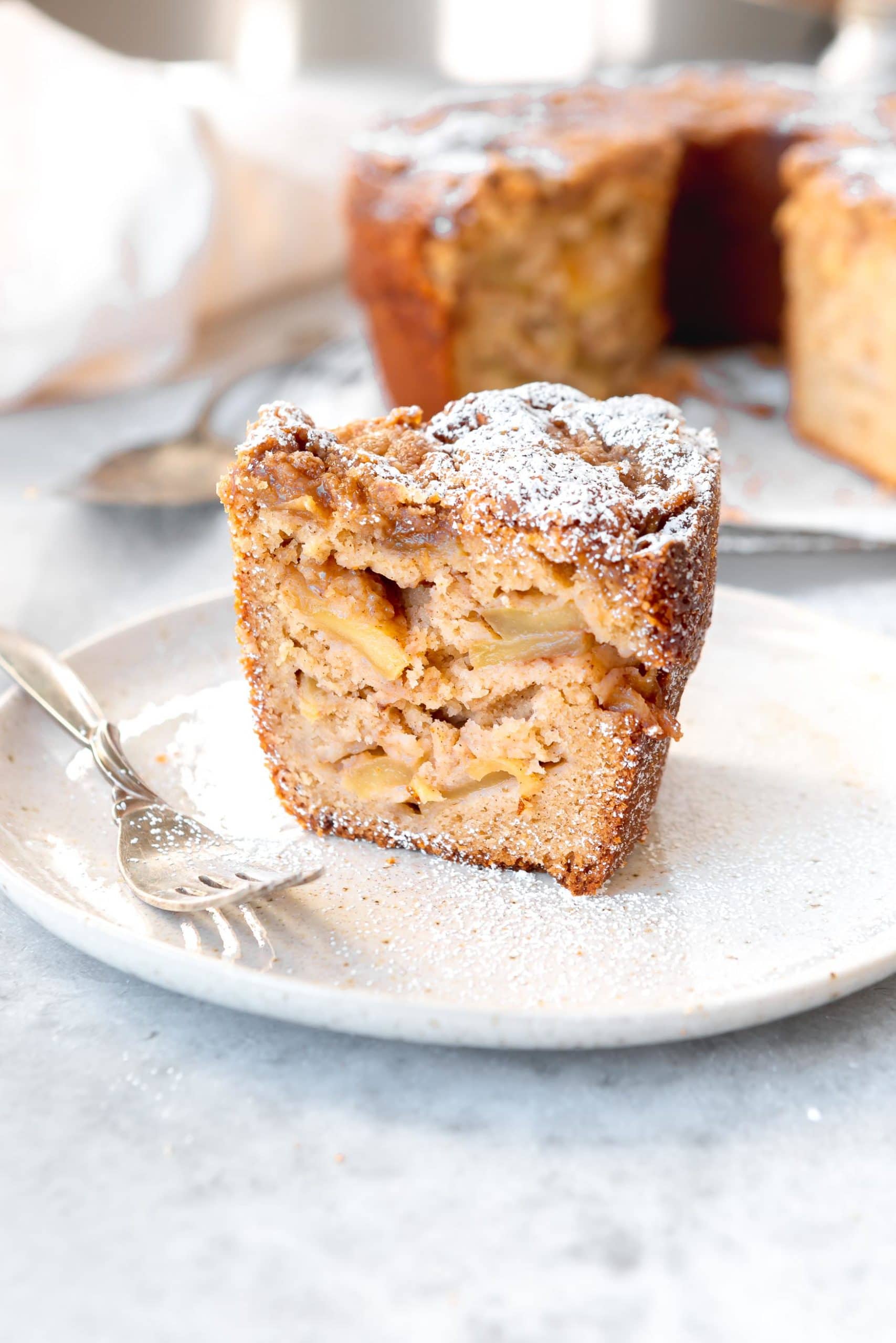 A slice of Jewish apple cake on a white dessert plate with a fork next to it. The cake is dense and filled with apple slices and there is powdered sugar on top.