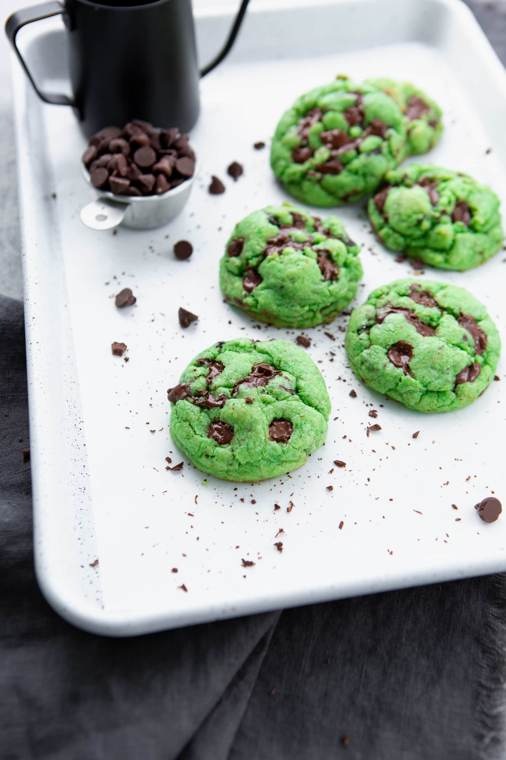 a photo of several green mint chocolate chip cookies on a parchment lined baking sheet with a cup of chocolate chips sitting next them.