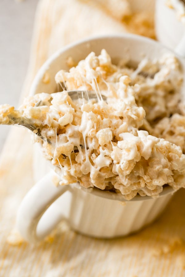A photo of a white mug full of gooey Rice Krispie treats with a spoonful being pulled out.