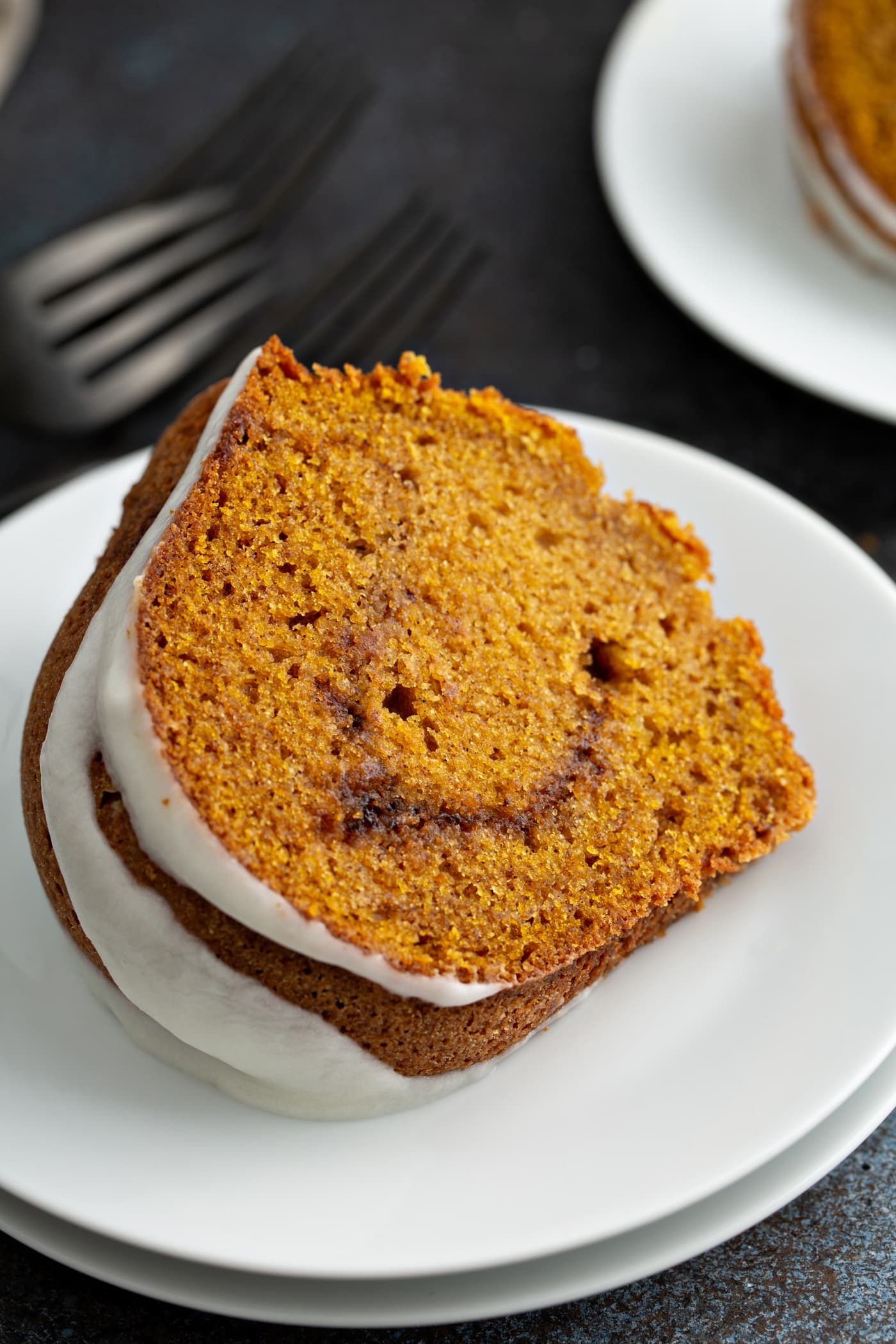 A slice of sour cream cinnamon swirl pumpkin cake on a dessert plate. The cake is drizzled with glaze and the cinnamon swirl in the middle is visible. A fork is next to the plate and another dessert plate is in the background.