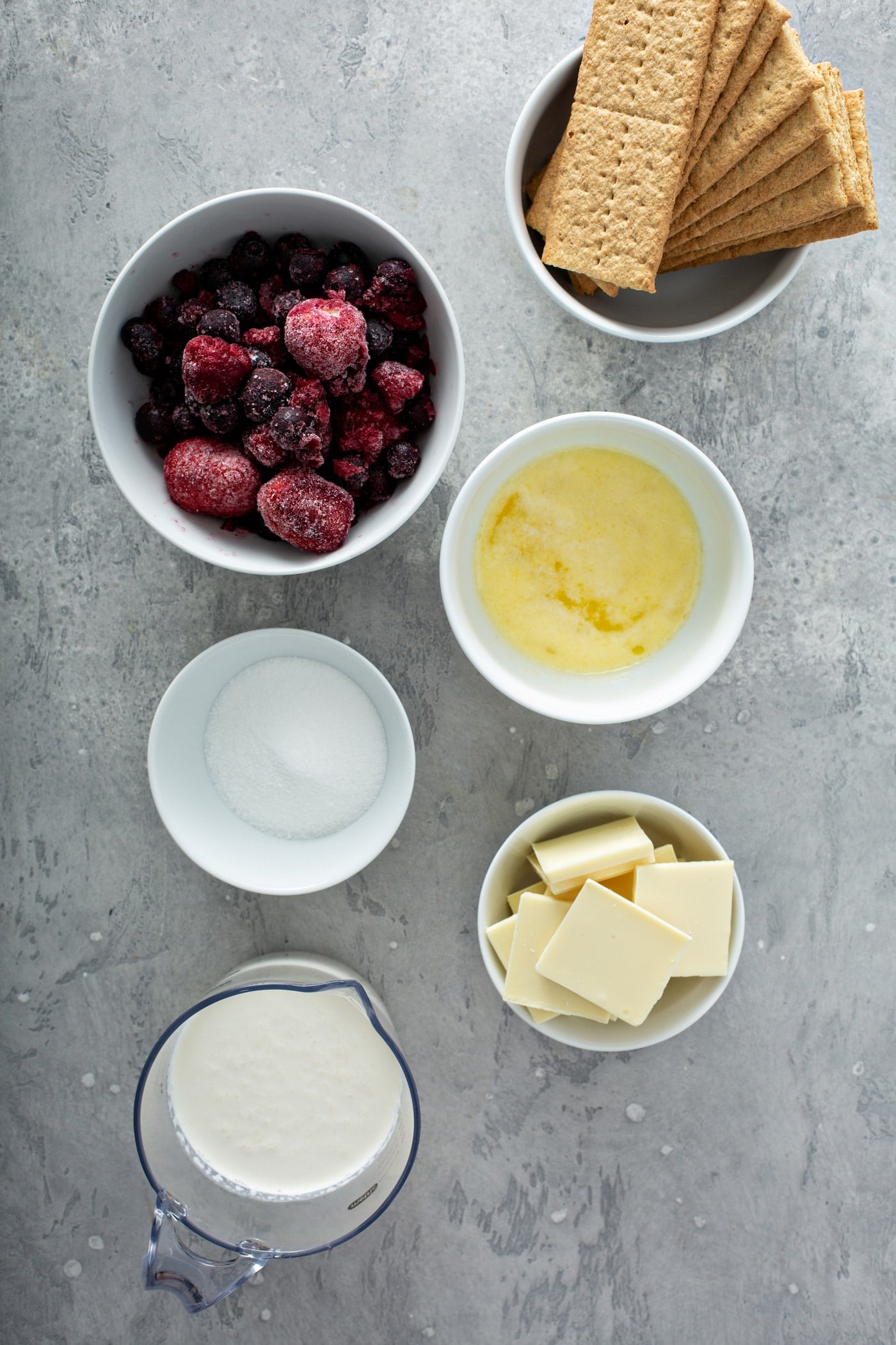 Small containers of ingredients for white chocolate berry pie. There are containers of butter, cubes of white chocolate, berries, graham crackers, sugar, and whipped cream.
