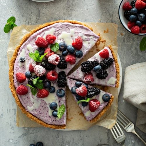 A berry white chocolate pie that has been sliced. A slice has been removed and two more slices are separated. The pie has a graham cracker crust and is topped with strawberries, blue berries and raspberries, with white chocolate sauce drizzled on top. A few mint leaves are scattered on top of the pie and on the table next to the pie. There are two forks, a napkin and a bowl of mixed berries on the table. A dessert plate with a slice of pie is in the background.