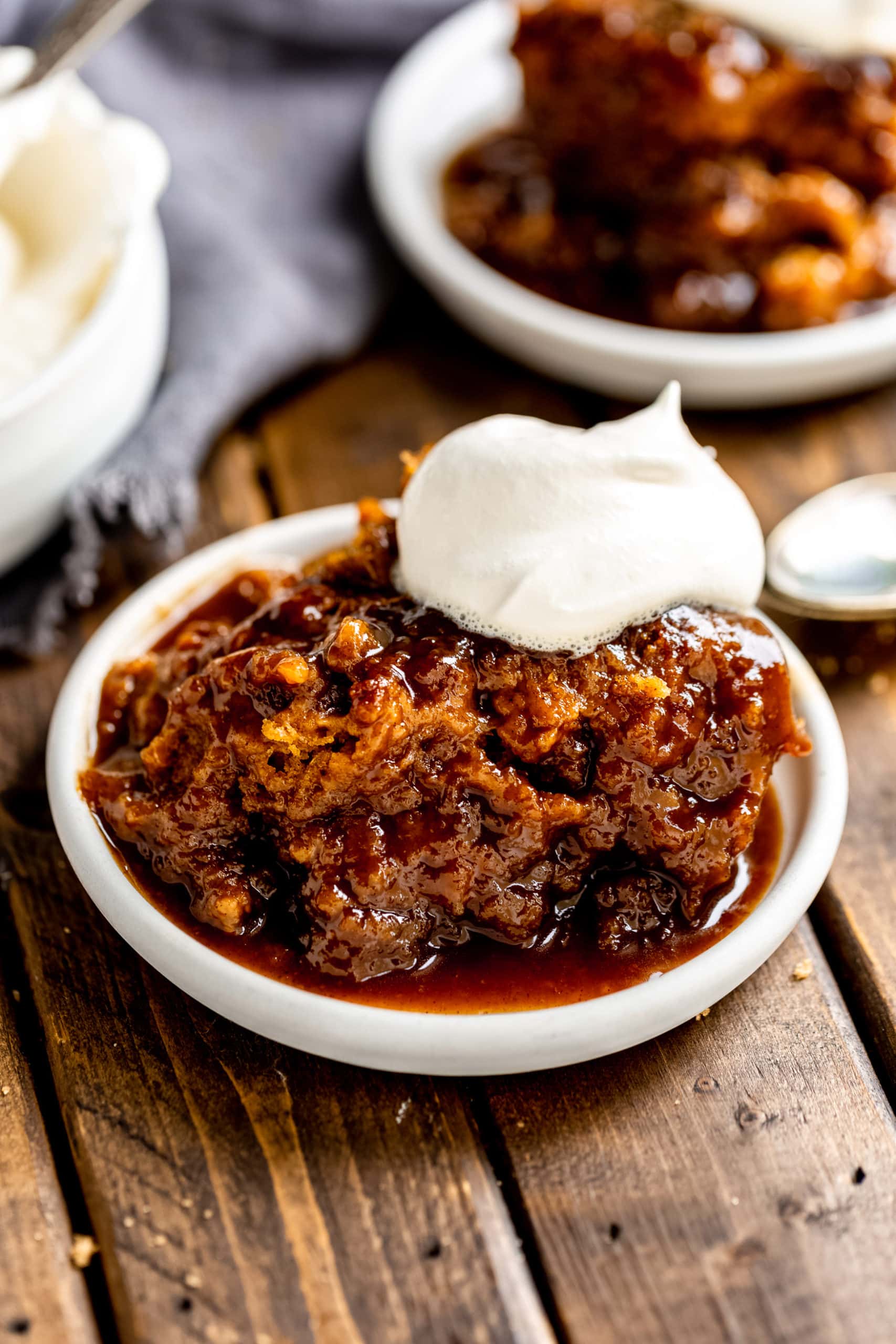 a photo of a serving of pumpkin lava cake in a small white saucer. the cake is sitting in a thick sauce that has seeping out of the cake and the cake is topped with whipped cream.