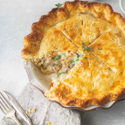A baked turkey pot pie with a slice removed. The crust is a golden brown and you can see the creamy filling with peas, carrots and turkey. Two forks are next to the pie.