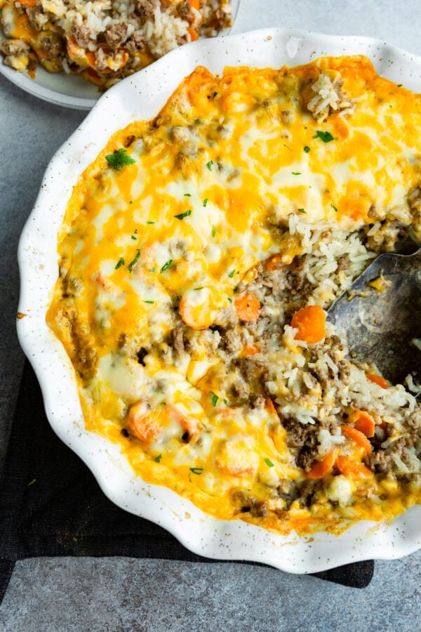 a photo of a round ceramic baking dish filled with a ground beef and rice casserole topped with melted cheese and a large serving spoon removing a scoop. a photo of a round ceramic baking dish filled with a ground beef and rice casserole topped with melted cheese and a large serving spoon removing a scoop.