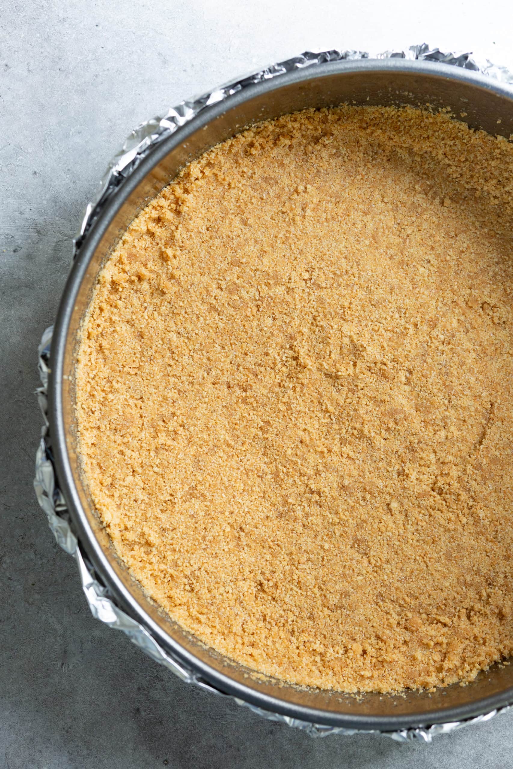 a photo of a graham cracker crust pressed down into a springform pan wrapped in foil ready to bake. a photo of a graham cracker crust pressed down into a springform pan wrapped in foil ready to bake.