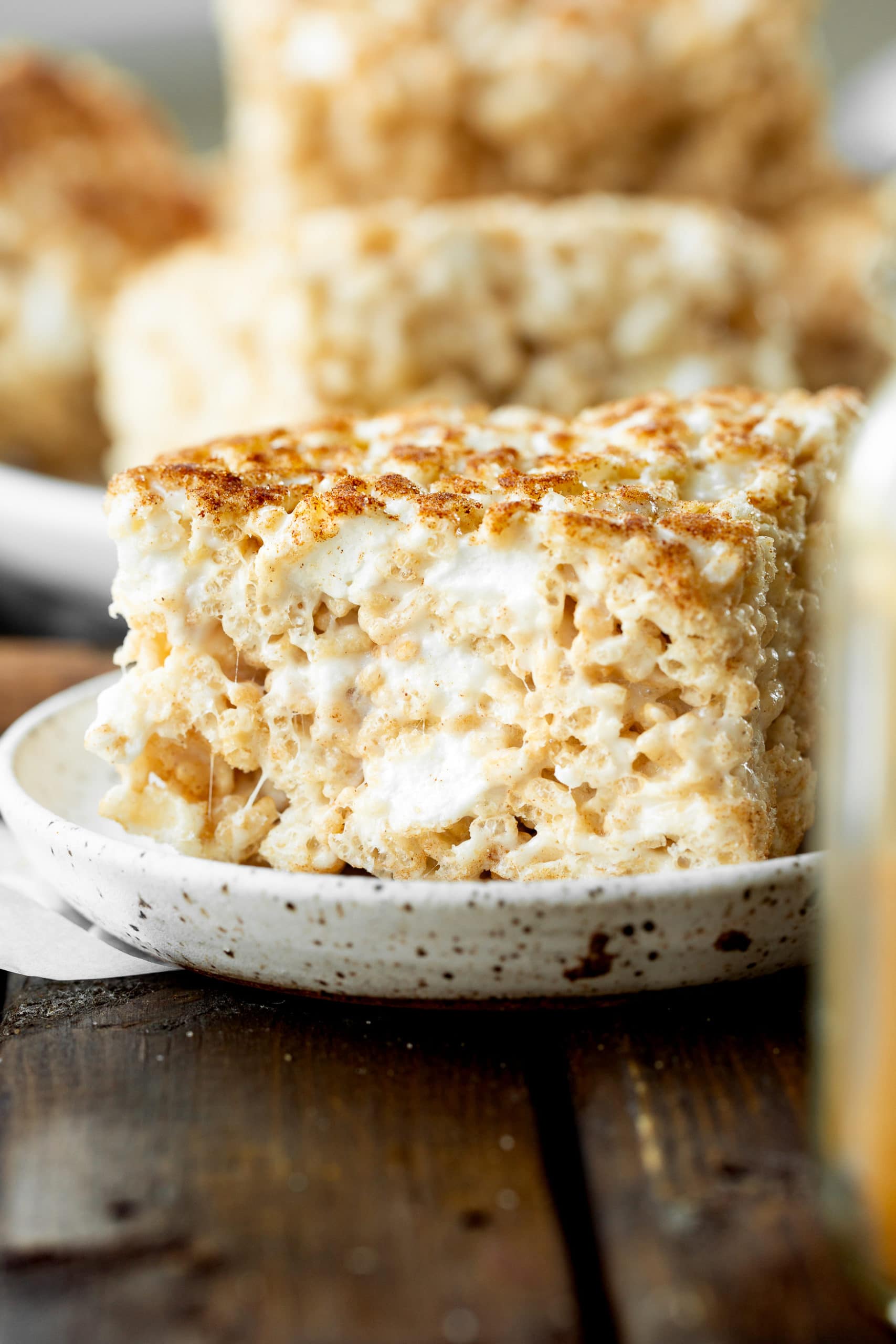 A photo of a single thick square churro rice krispie treat sitting on a small saucer and the treat is full of gooey marshmallows and topped with cinnamon sugar. A photo of a single thick square churro rice krispie treat sitting on a small saucer and the treat is full of gooey marshmallows and topped with cinnamon sugar.