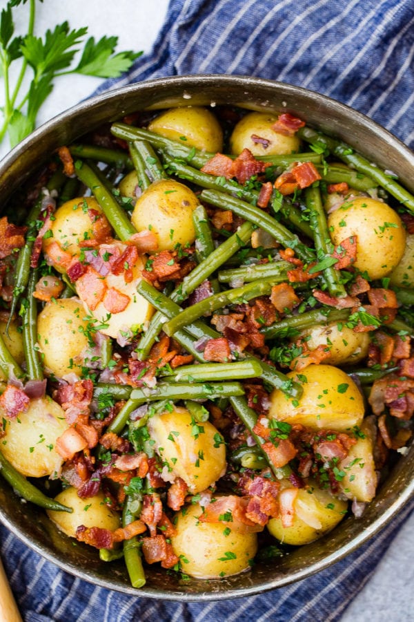 A photo of a silver serving dish sitting on a blue cloth napkin that is full of cooked fresh green beans, potatoes, and bacon pieces sprinkled with fresh parsley. A photo of a silver serving dish sitting on a blue cloth napkin that is full of cooked fresh green beans, potatoes, and bacon pieces sprinkled with fresh parsley.