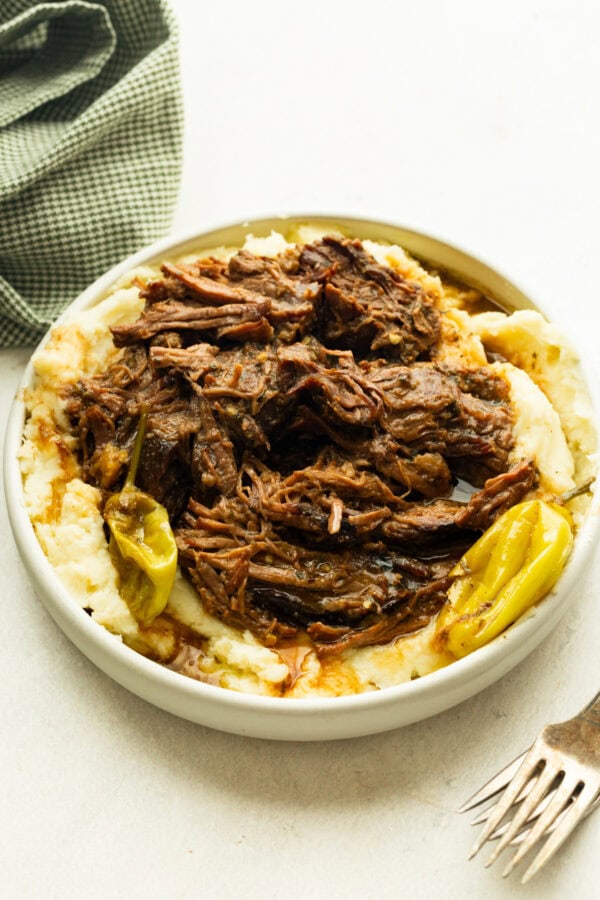 a large serving bowl full of juicy, tender beef served over the top of creamy mashed potatoes and whole pepperoncicnis around the bowl a large serving bowl full of juicy, tender beef served over the top of creamy mashed potatoes and whole pepperoncicnis around the bowl