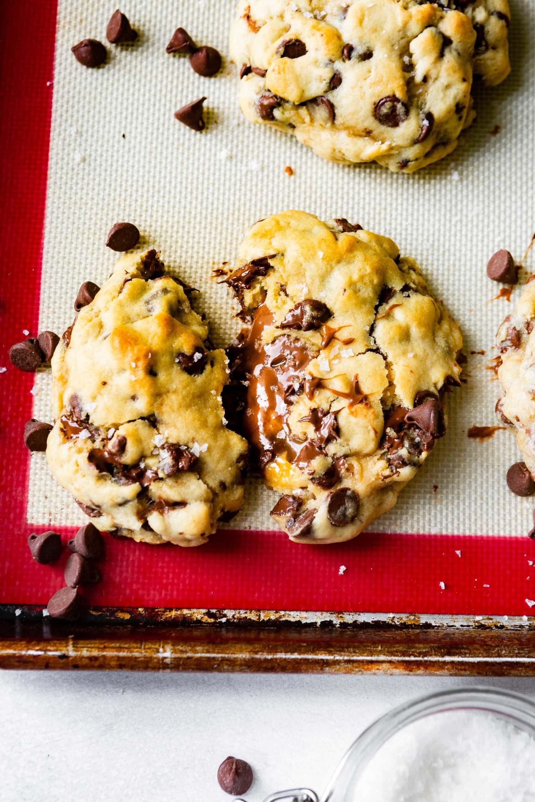 a thick chocolate chip cookie stuffed with nutella and dulce de leche caramel split in half with chocolate chips sprinkled around it a thick chocolate chip cookie stuffed with nutella and dulce de leche caramel split in half with chocolate chips sprinkled around it