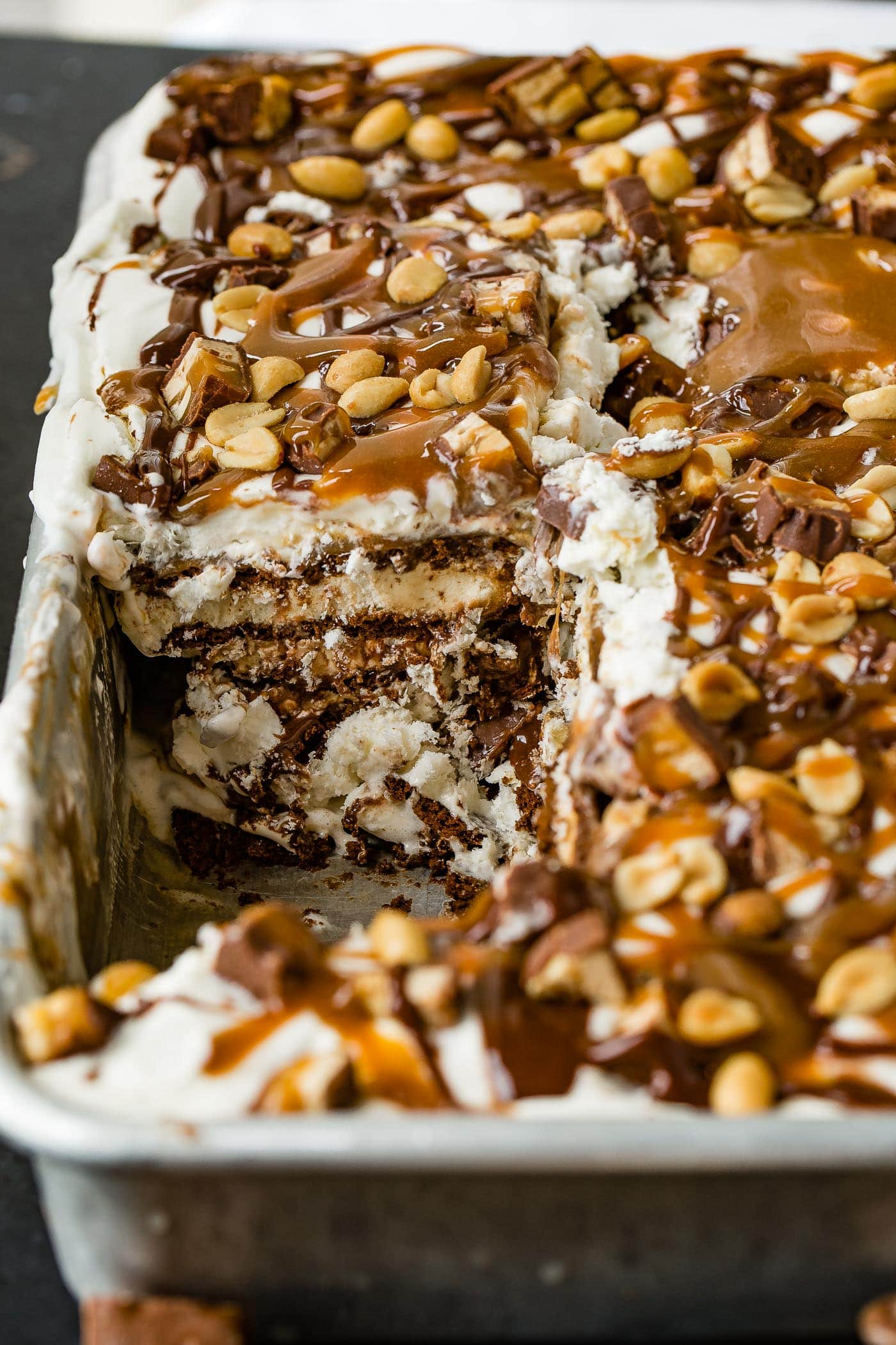 A pan of snickers ice cream cake with a piece removed. There are layers of ice cream, cake, fudge, caramel, whipped cream and candy bars. The top is drizzled with hot fudge, caramel topping and salted peanuts.