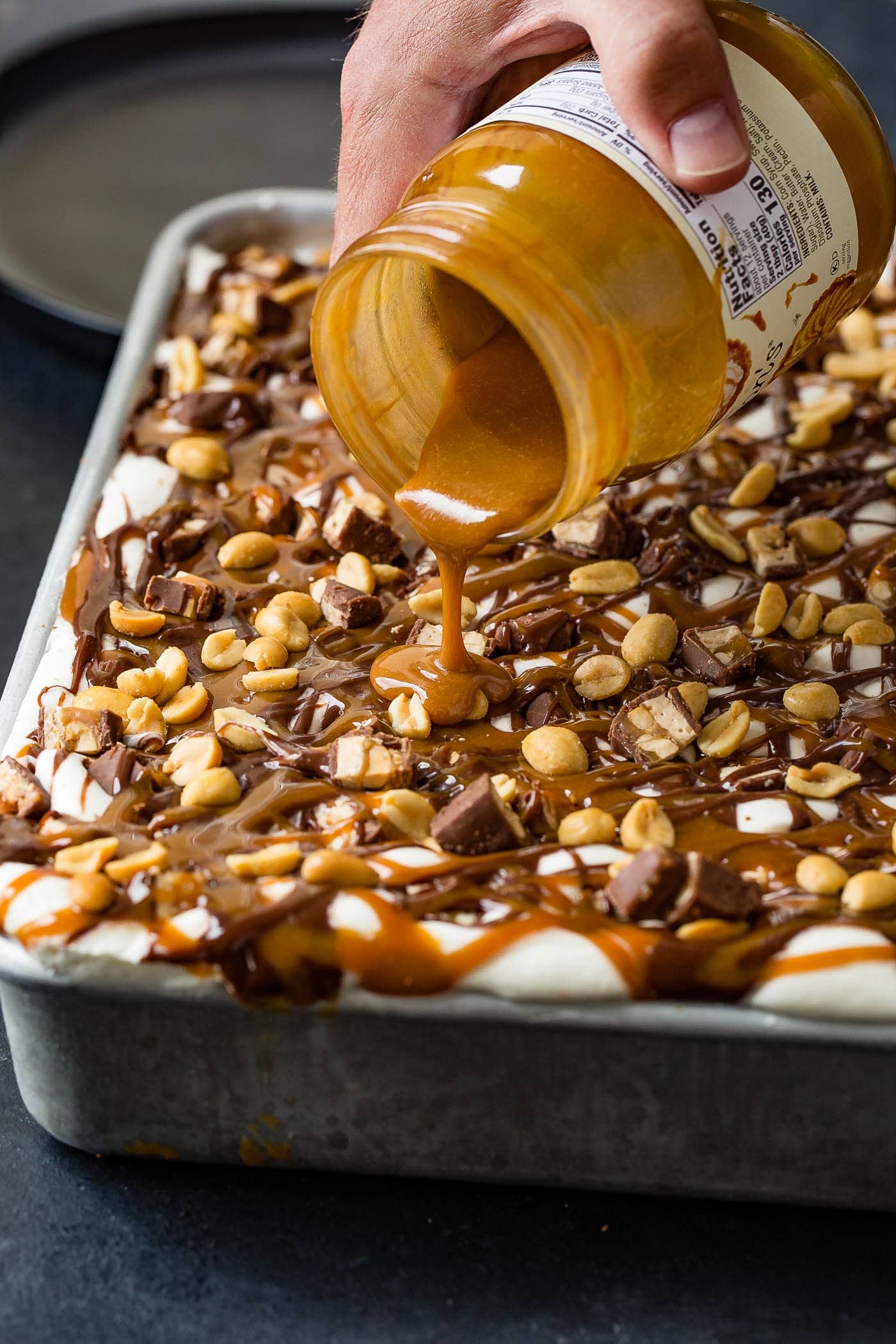 A jar of melted caramel topping being poured over the top of a snickers ice cream cake. The top of the cake is covered with whipped cream, drizzles of hot fudge, and salted peanuts.
