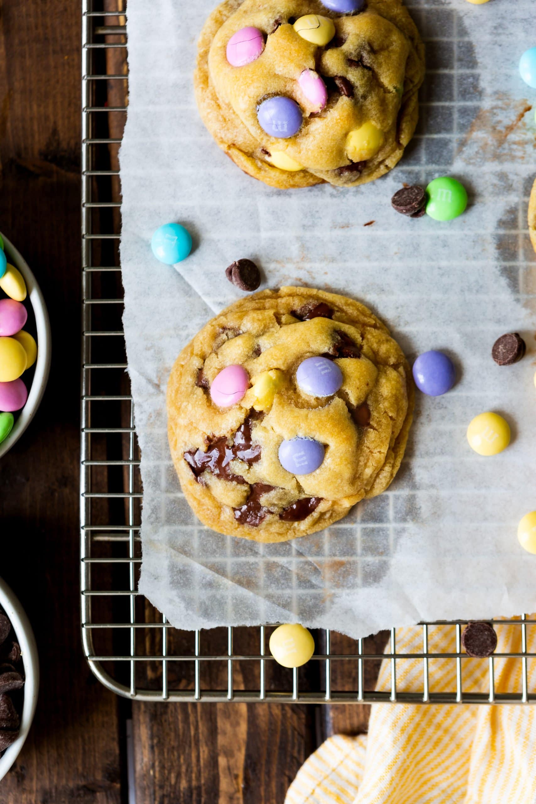 a photo of single thick and chewy perfectly golden Easter m&m chocolate chip cookie sitting on a piece of parchment paper on top of a wire cooling rack with a few m&m's and chocolate chips scattered around it. a photo of single thick and chewy perfectly golden Easter m&m chocolate chip cookie sitting on a piece of parchment paper on top of a wire cooling rack with a few m&m's and chocolate chips scattered around it.