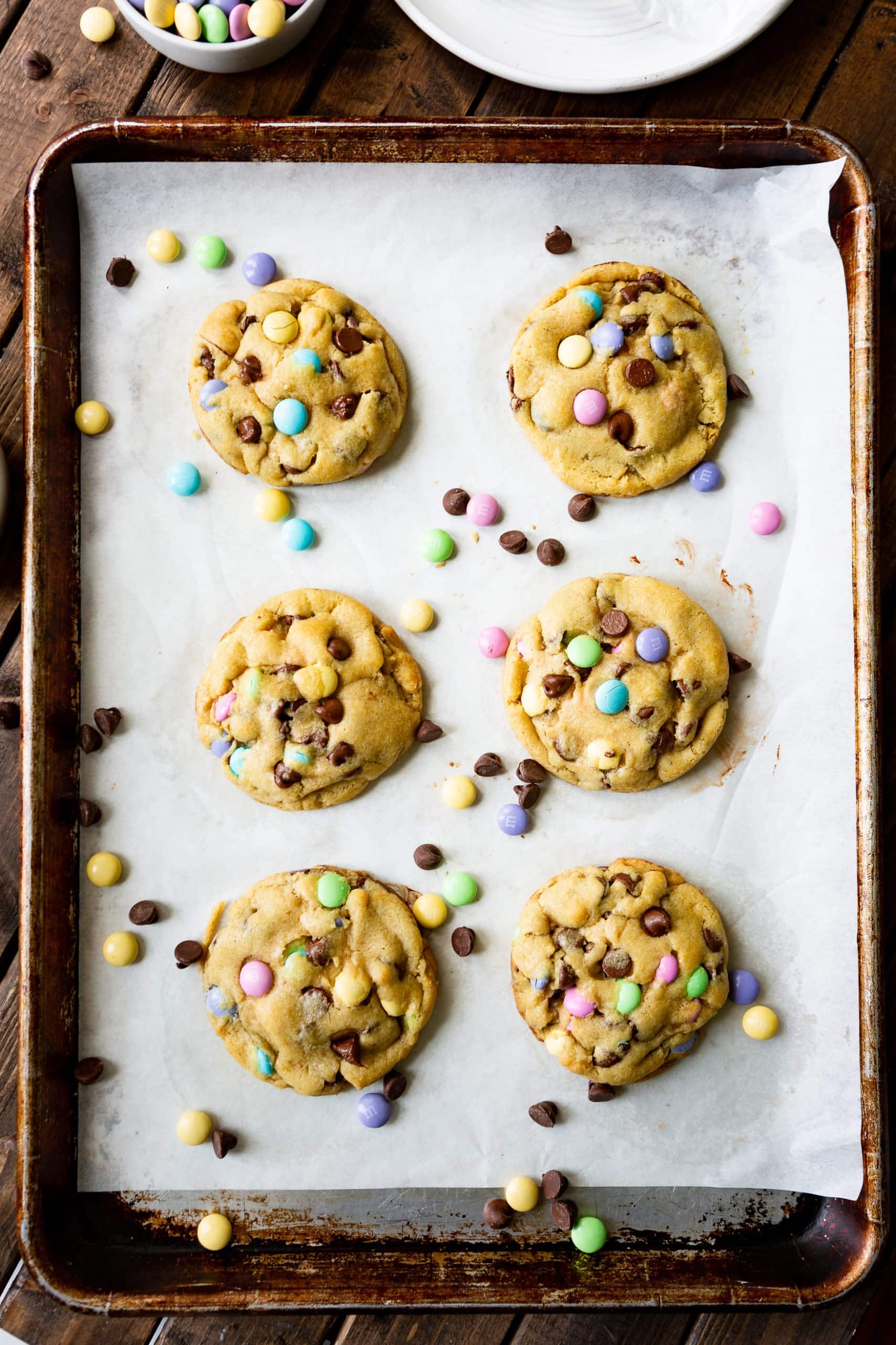 a photo of six large Easter m&m chocolate chip cookies sitting on a baking sheet lined with parchment paper with chocolate chips and m&m's scattered around them. a photo of six large Easter m&m chocolate chip cookies sitting on a baking sheet lined with parchment paper with chocolate chips and m&m's scattered around them.