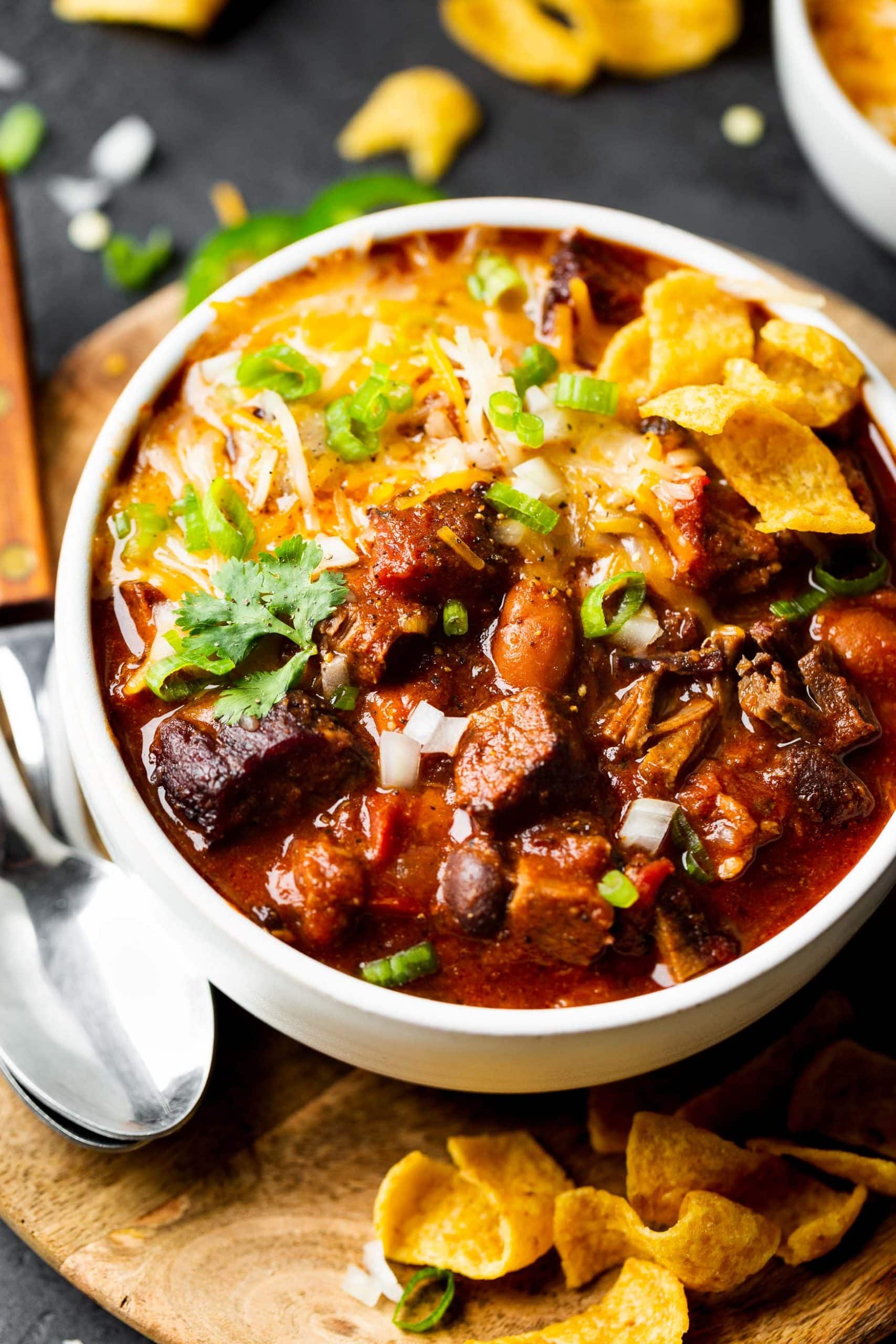 a close up photo of a white soup bowl full of homemade brisket chili topped with fritos, melting shredded cheese and slices of green onions. a close up photo of a white soup bowl full of homemade brisket chili topped with fritos, melting shredded cheese and slices of green onions.