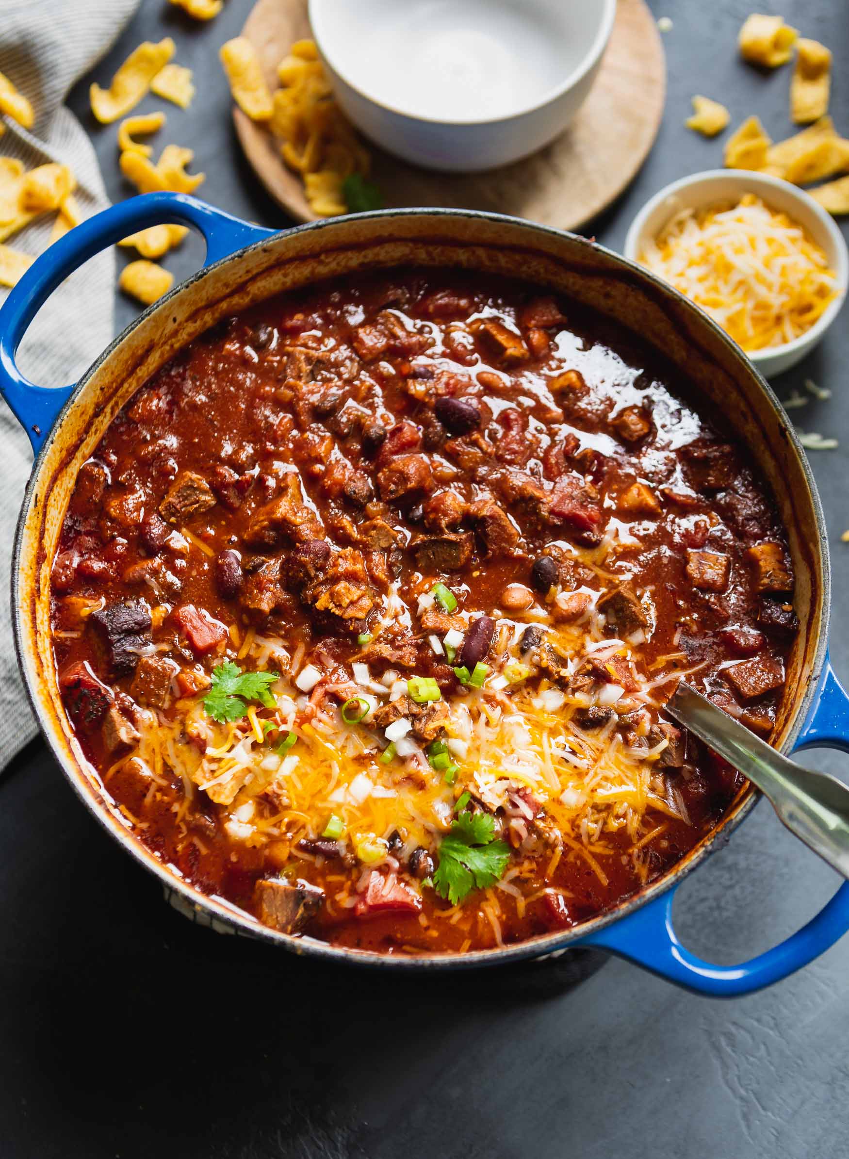 a photo of a large dutch oven full of brisket chili topped with melting shredded cheese and slice green onions with a large silver serving spoon resting on the edge of the pot. a photo of a large dutch oven full of brisket chili topped with melting shredded cheese and slice green onions with a large silver serving spoon resting on the edge of the pot.
