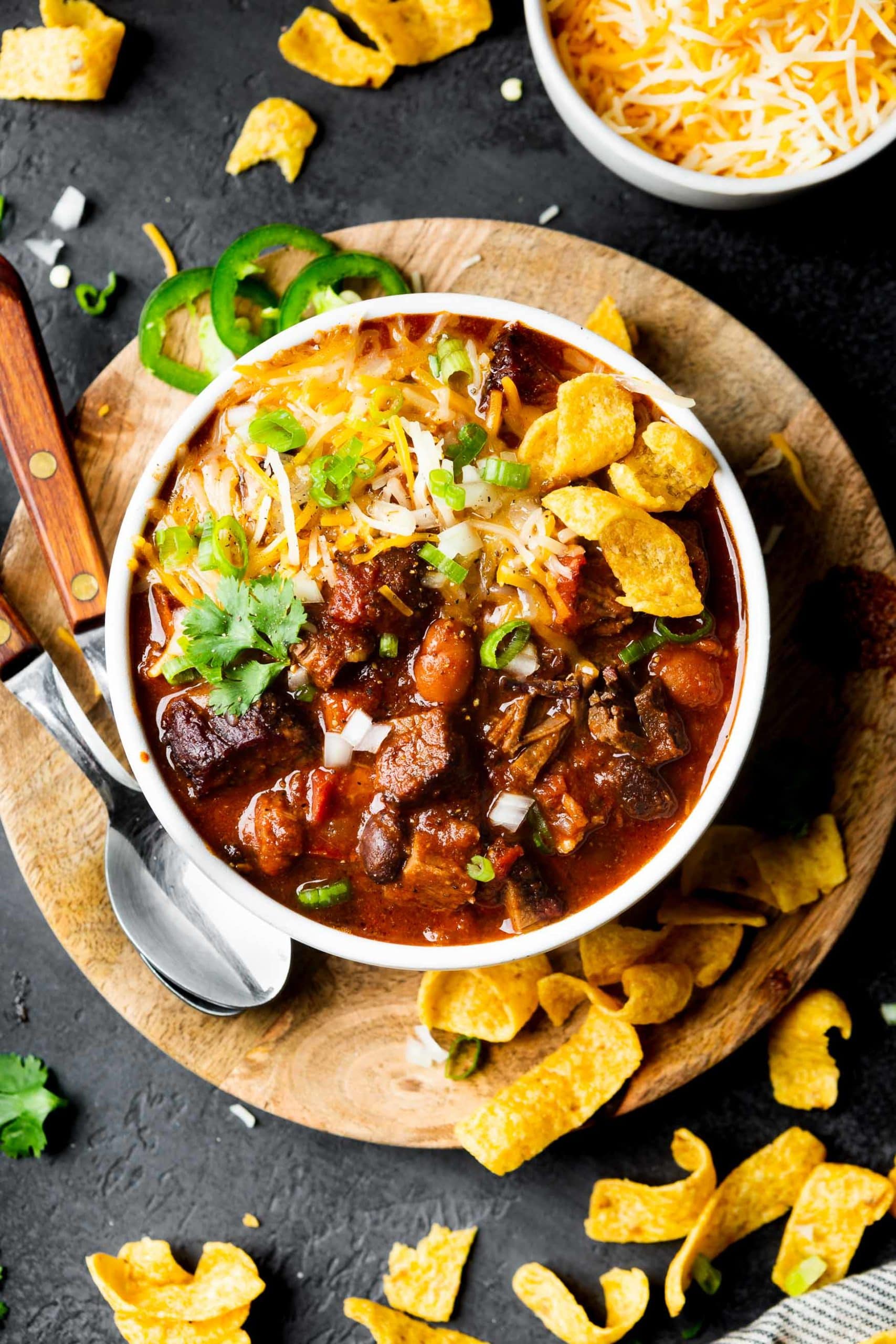 a photo of a bowl of brisket chili sitting on a circular wooden charger with two spoons sitting next to the bowl, fritos scattered around, and slices of fresh jalapeno. a photo of a bowl of brisket chili sitting on a circular wooden charger with two spoons sitting next to the bowl, fritos scattered around, and slices of fresh jalapeno.