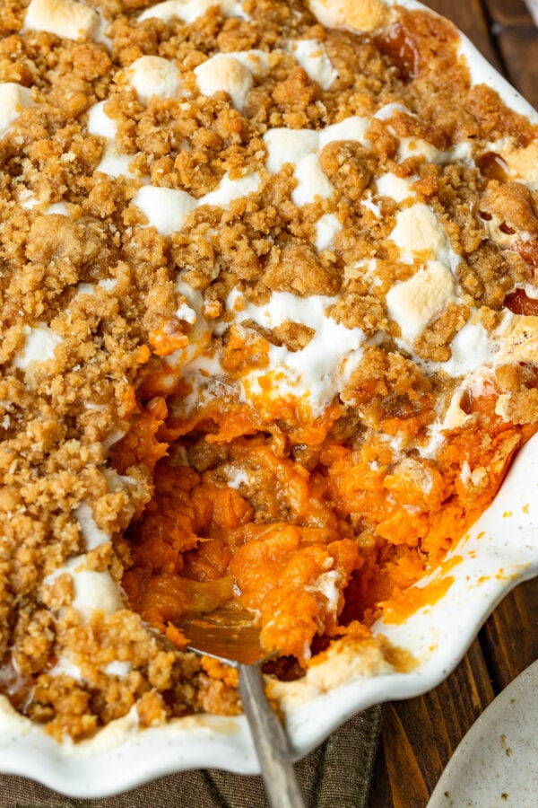 a photo of a round baking dish full of sweet potato casserole topped with gooey marshmallows and a brown sugar streusel topping with a serving being scooped out of the dish a photo of a round baking dish full of sweet potato casserole topped with gooey marshmallows and a brown sugar streusel topping with a serving being scooped out of the dish