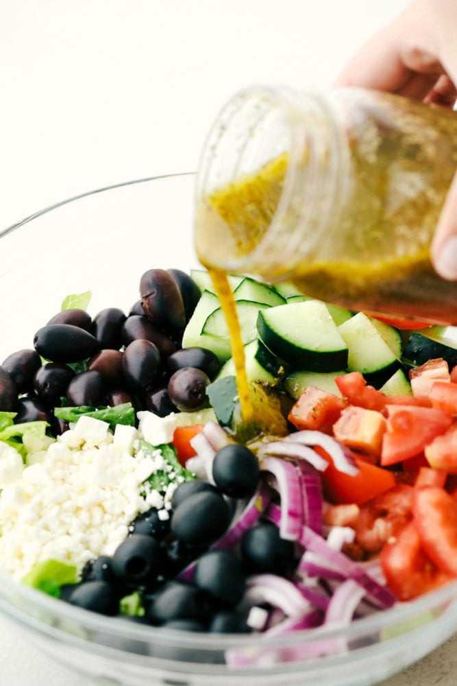 Greek Salad with olives, feta cheese, tomatoes and cucumbers with Greek Salad Dressing being poured over top. 